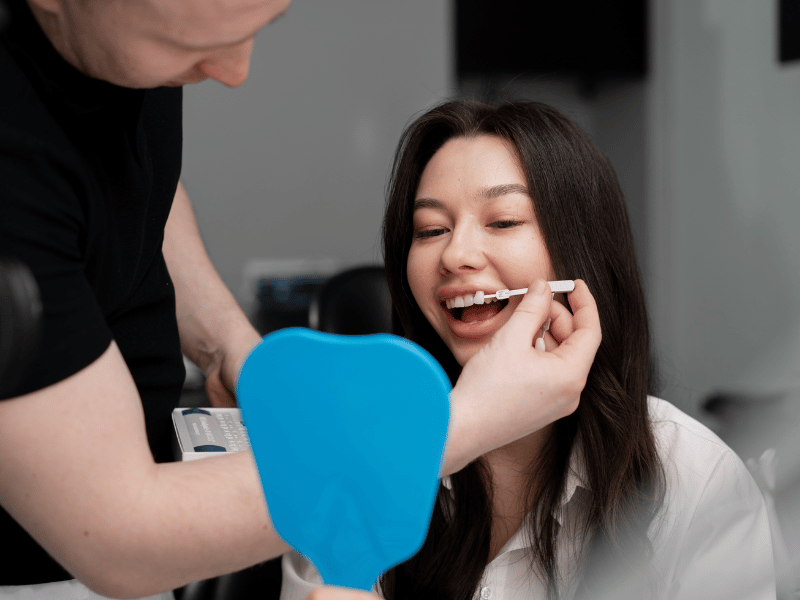 Zirconium Implants Dentist holding a tooth shade guide beside a smiling woman's teeth while she looks into a blue hand mirror